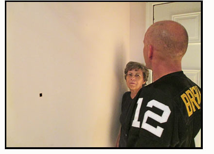 Photo shows the man in the football jersey looking toward his right about head-high at the plain light-colored wall.  On the wall toward his left about chest-high is a black spot.  Dona is watching him scan.